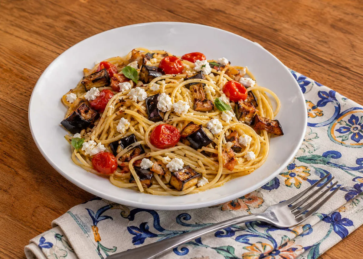 Spaghetti with roasted eggplant, cherry tomatoes and crumbled feta on a white plate over a patterned napkin